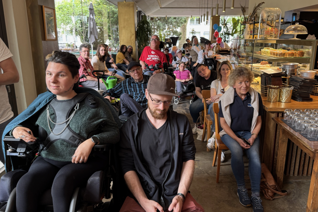 Größere Gruppe von Menschen mit und ohne sichtbare Behinderungen sitzt eng gedrängt in einem Café mit Tischen, Stühlen und einer Theke mit Kuchen und Gläsern