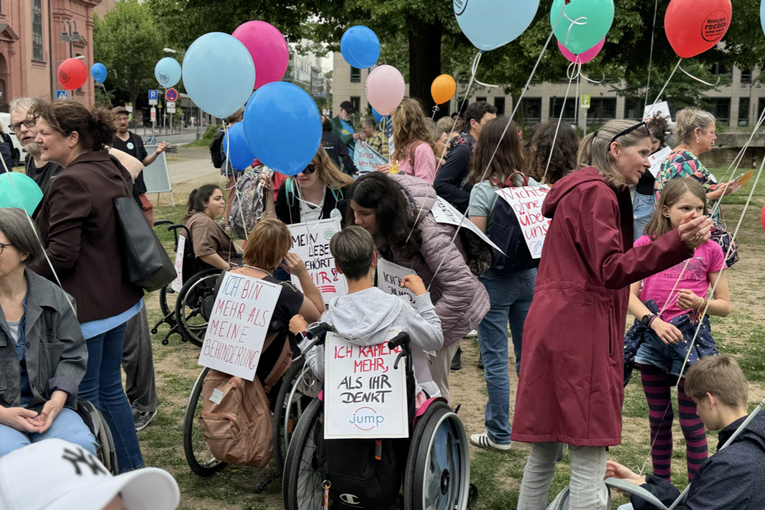 Menschen auf einer Wiese bei der Mahnwache mit bunten Luftballons. Einige Personen tragen Schilder. Eine Person im Rollstuhl trägt ein Schild mit der Aufschrift 'ICH KANN MEHR ALS IHR DENKT'