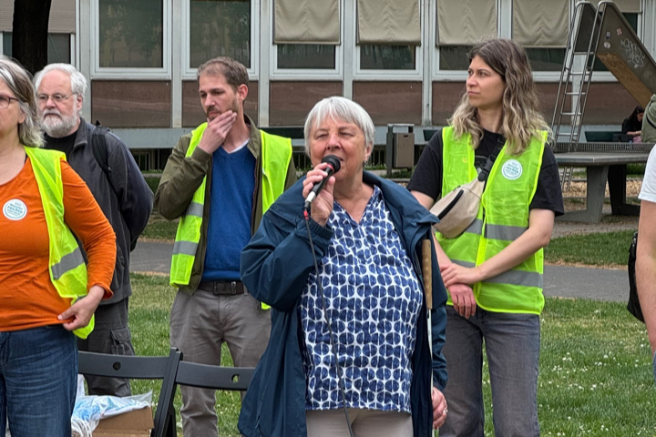 Fünf Personen, von denen drei gelbe Warnwesten tragen, stehen auf einer Wiese, eine grauhaarige Frau spricht in ein Mikrofon