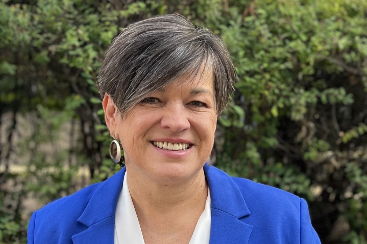 Portait einer Frau mittleren Alters mit grauem Kurzhaarschnitt, großen Ohrringen und einem intensiv blauen Blazer. Im Hintergrund sind Bäume.