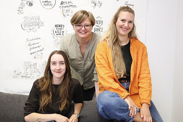 Gruppenbild: 3 Frauen auf und hinter einem Sofa vor einem beschrifteten Whiteboard.