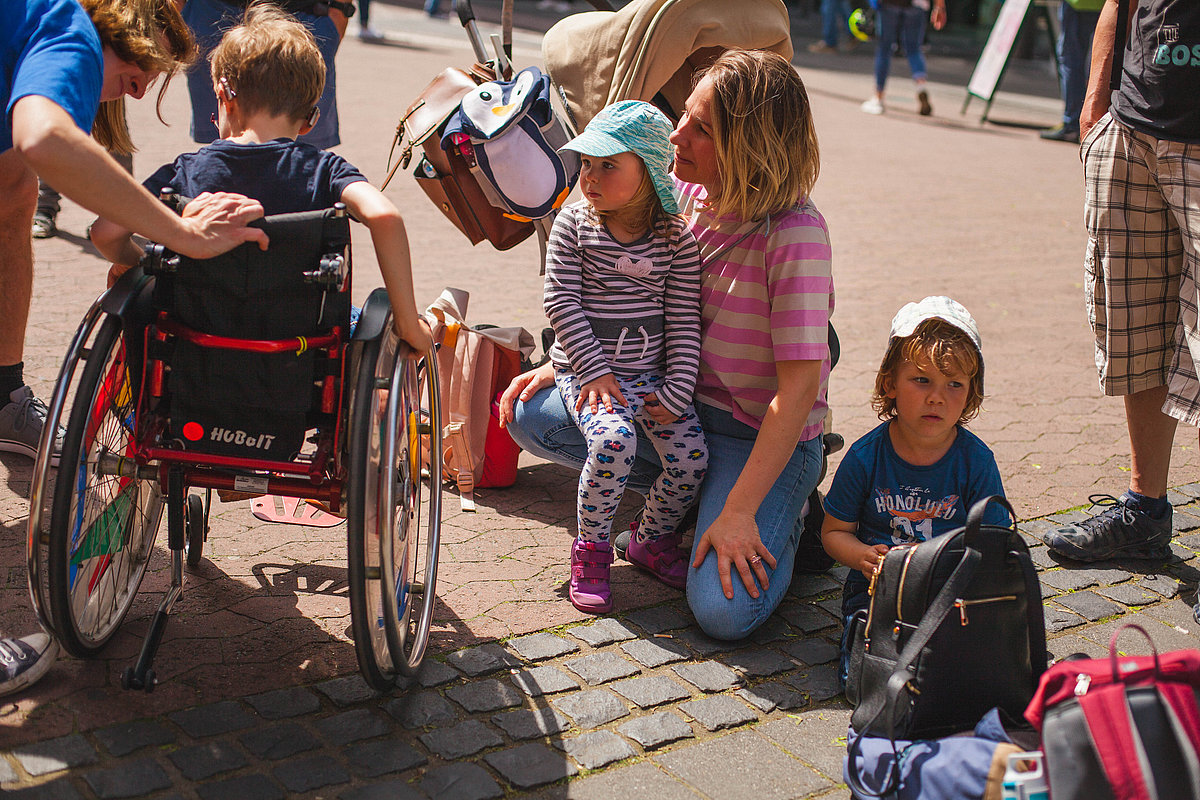 Eine Frau sitzt umgeben von Taschen mit zwei Kindern auf dem Boden. Daneben ist ein Junge im Rollstuhl.
