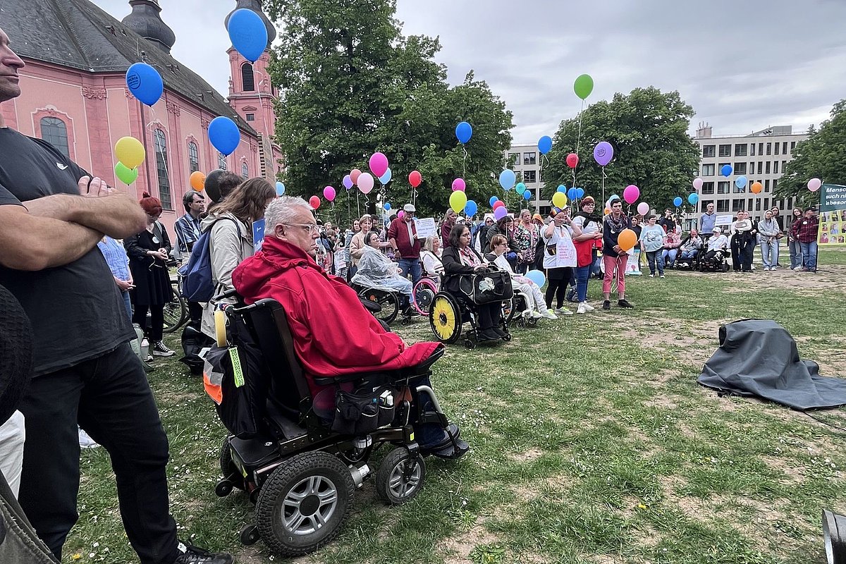Menschen mit und ohne Rollstuhl versammeln sich auf einer Wiese vor einem Gebäude mit rosa Fassade und halten bunte Luftballons