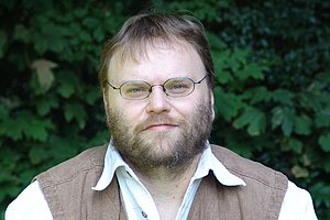 Porträt eines Mannes mit Brille und Bart vor einer grünen Hecke. Er trägt ein weißes Hemd und eine blassbraune Weste.