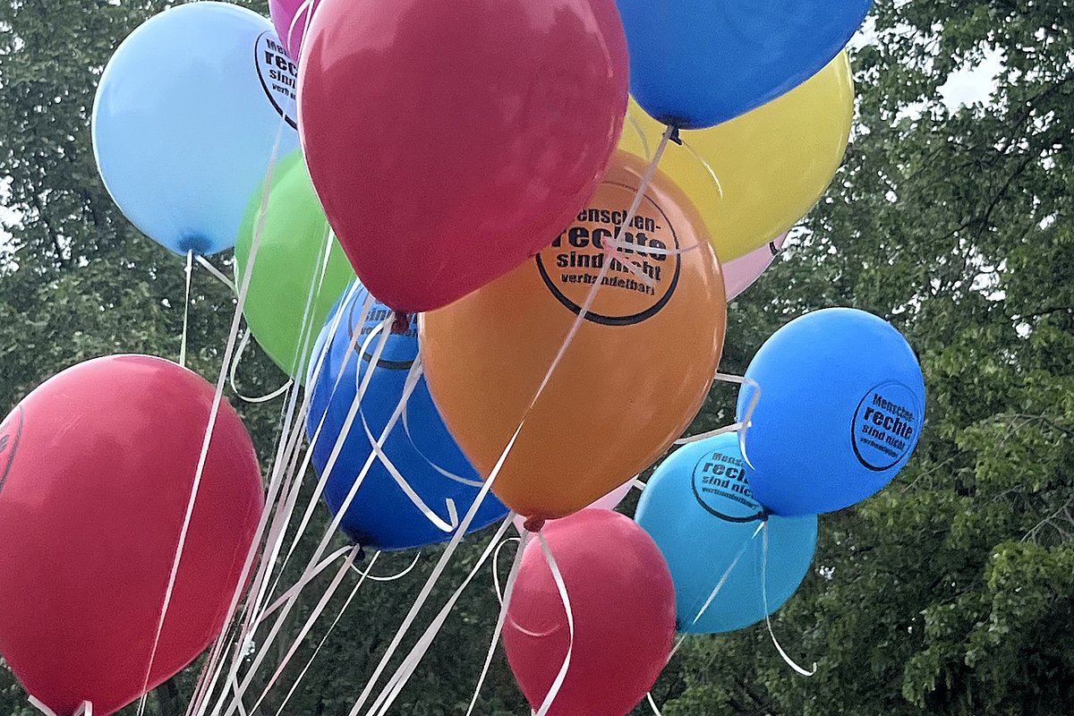 Mehrere bunte Luftballons mit dem Aufdruck 'Menschenrechte sind nicht verhandelbar' schweben vor grünen Bäumen
