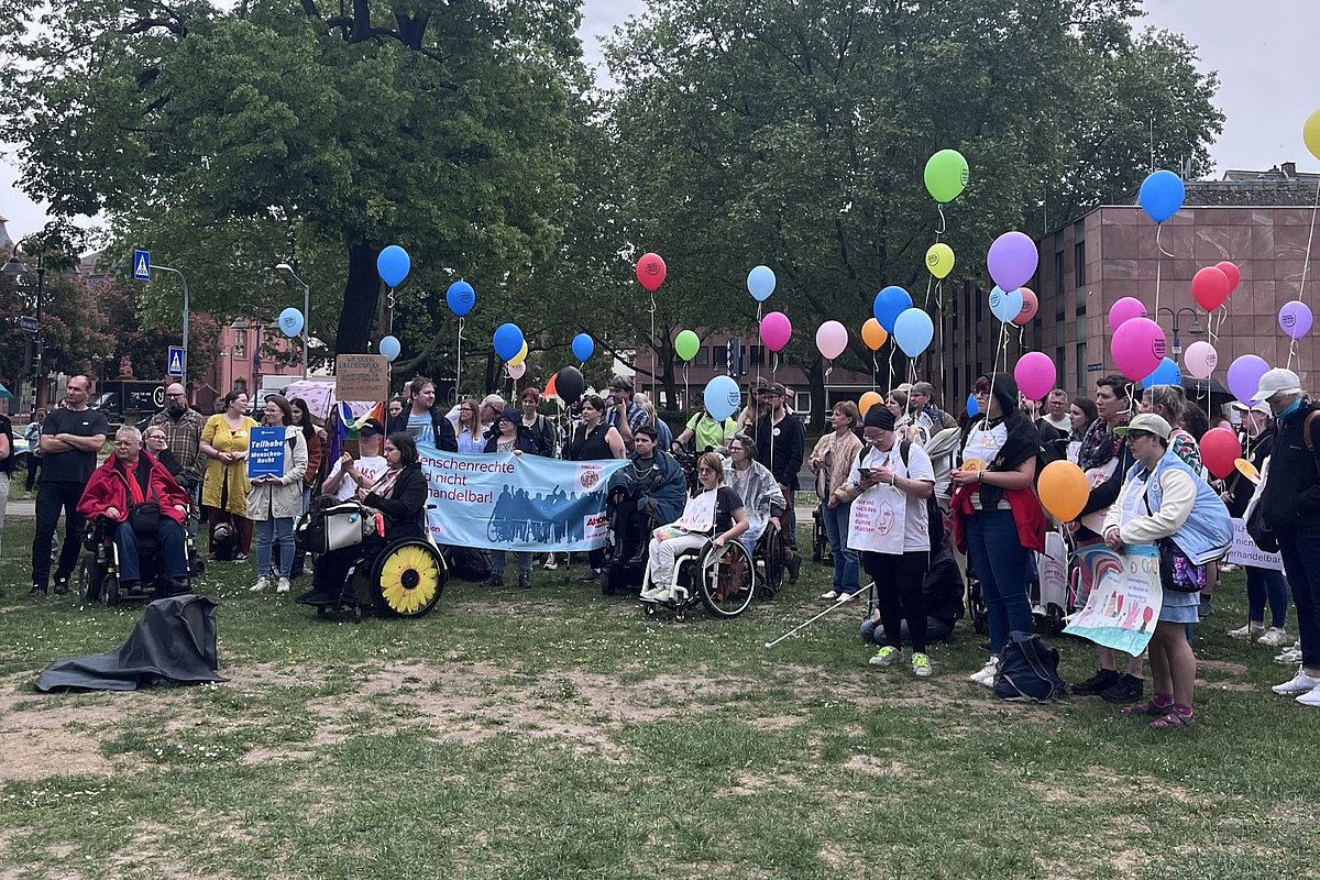 Gruppe von Menschen mit bunten Luftballons und Transparenten auf einer Wiese vor Bäumen und einem Gebäude