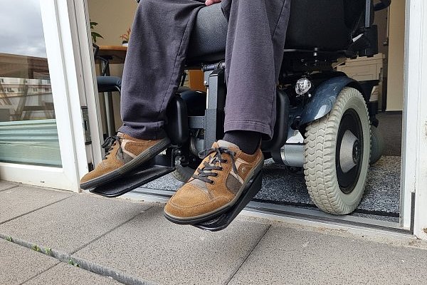 Nahaufnahme auf die Beine einer Person im Rollstuhl, die versucht, eine Türschwelle zu überwinden.