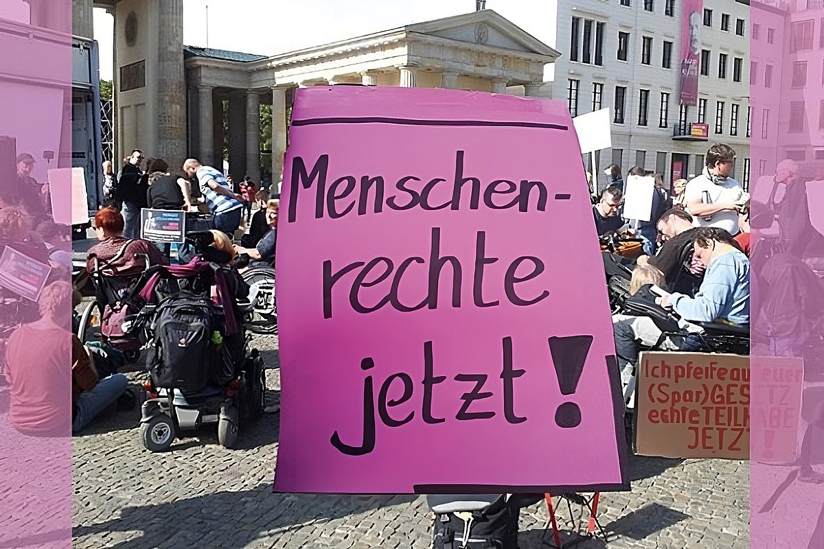 Eine Gruppe von Demonstrierenden mit und ohne sichtbare Behinderung. Im Vordergrund ein pinkes Schild mit der Aufschrift "Menschenrechte jetzt!".