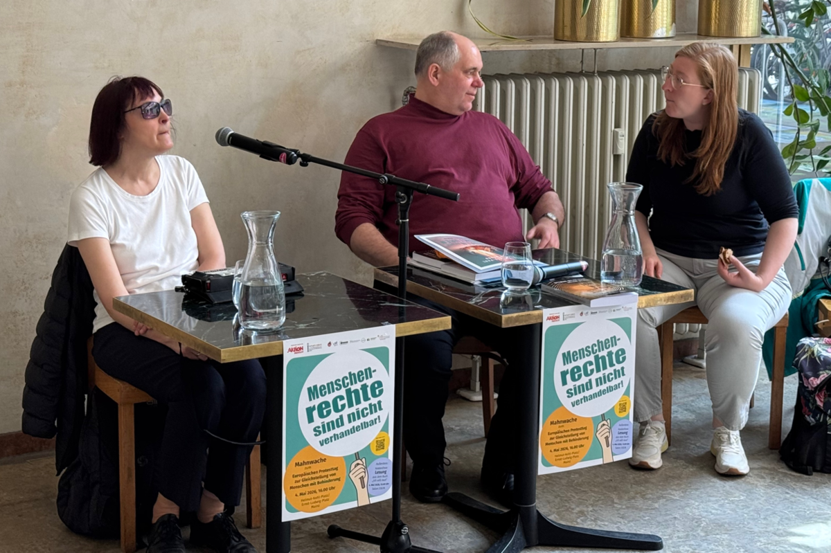 Drei Personen sitzen an zwei Tischen mit Wasserkrügen und Gläsern, an den Tischen hängen Plakate mit der Aufschrift 'Menschenrechte sind nicht verhandelbar'