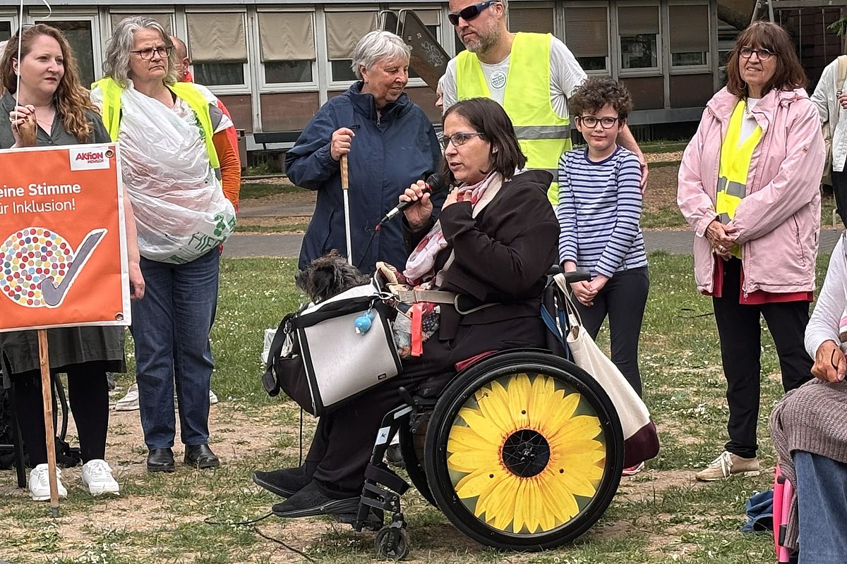 Person im Rollstuhl mit Sonnenblumenmotiv auf den Rädern spricht in ein Mikrofon vor einer Gruppe von Menschen, einige tragen gelbe Warnwesten