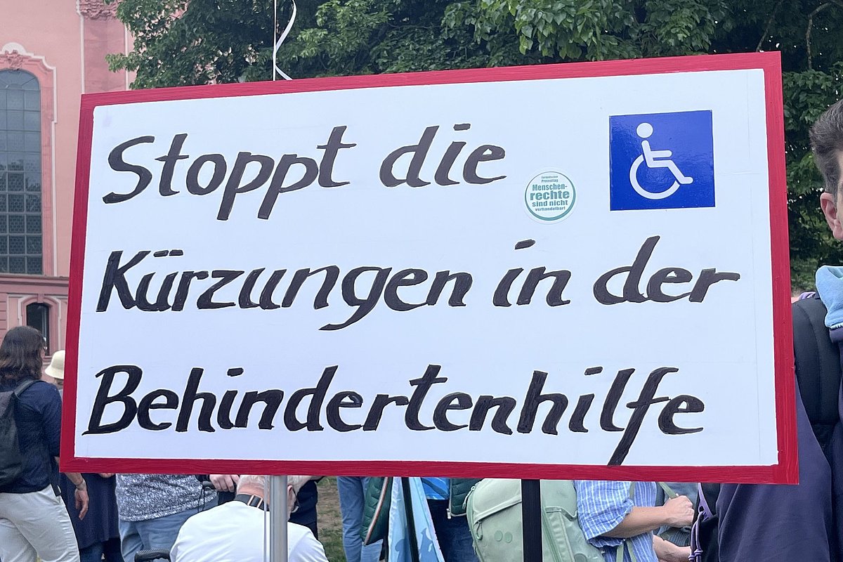 Schild mit der Aufschrift 'Stoppt die Kürzungen in der Behindertenhilfe' und einem Rollstuhlsymbol