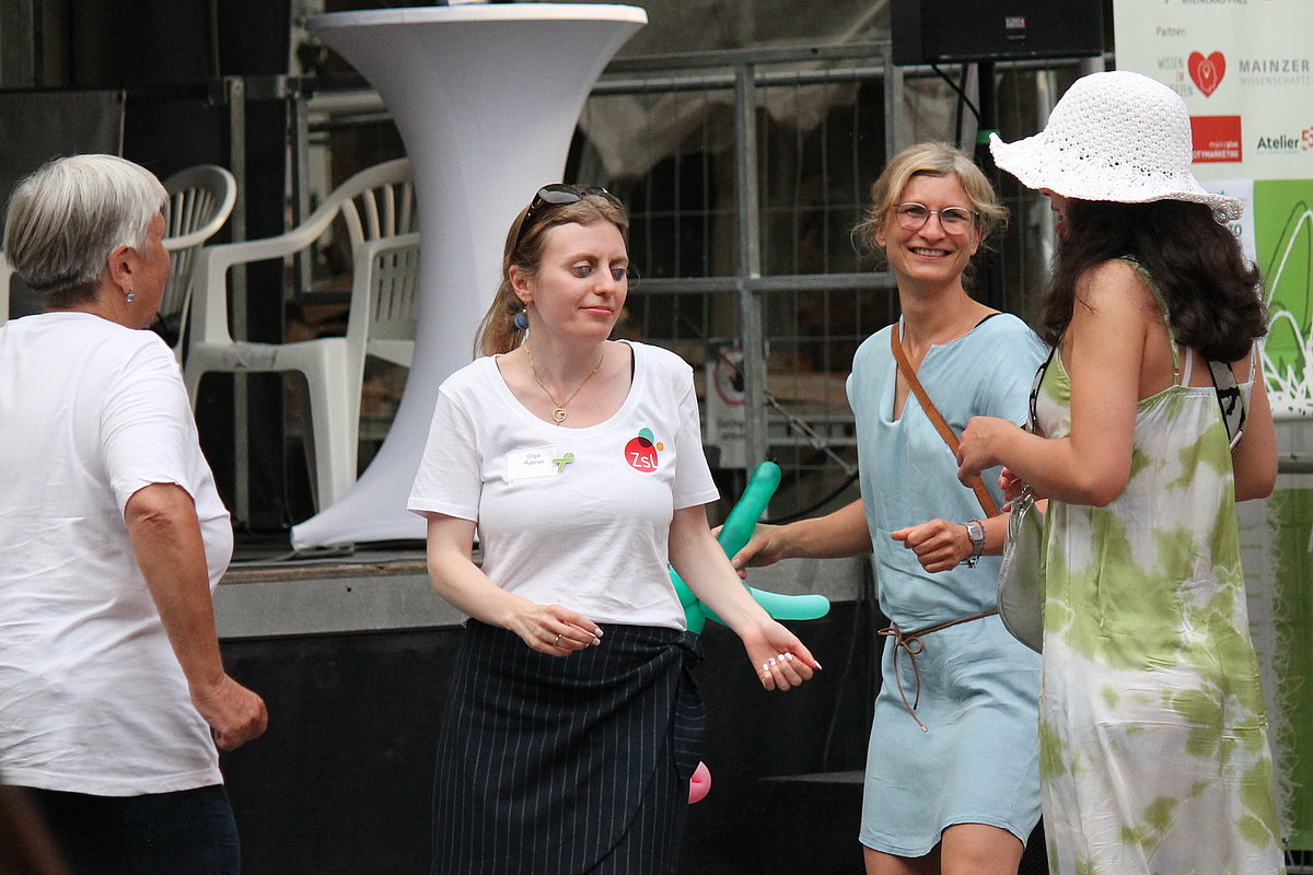 Vier Frauen tanzen. Eine davon trägt einen großen weißen Hut.  