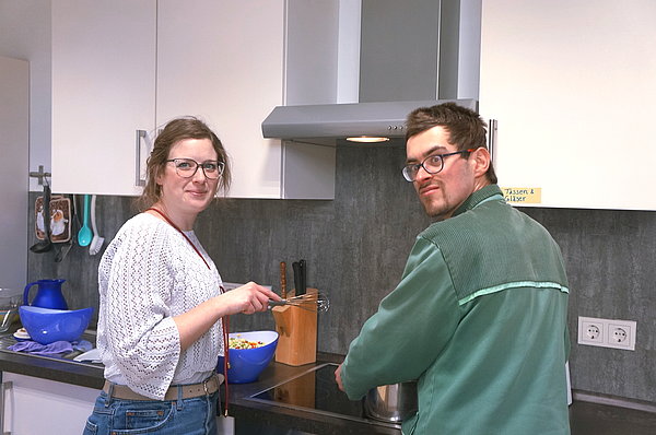 A woman and a man are cooking at a stove. They look over their shoulders into the camera.